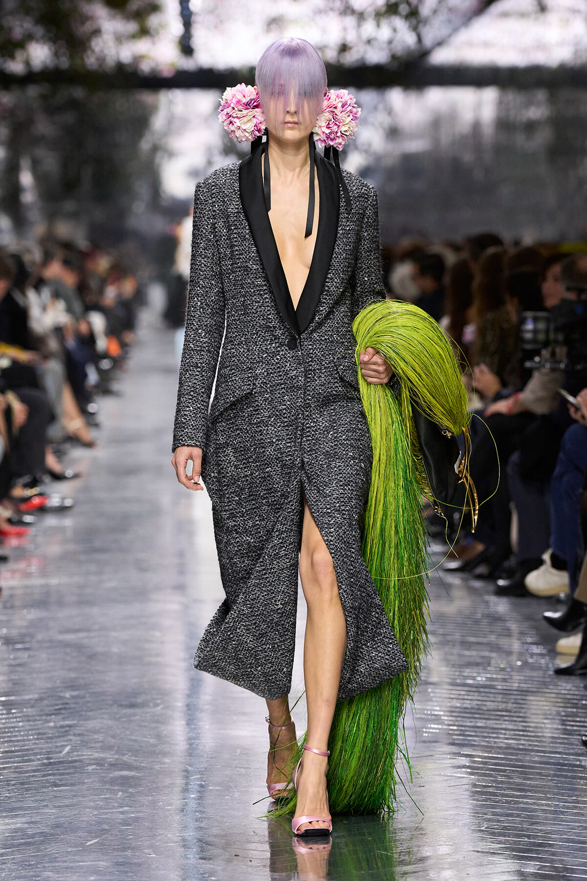 Runway model in a textured gray coat with black lapels and deep V, pink heels, and a bright green fringe prop; lavender hair with pink floral headpiece