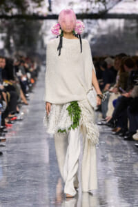 Runway model in an oversized cream knit poncho with a pink bob and large pink flower headpieces.