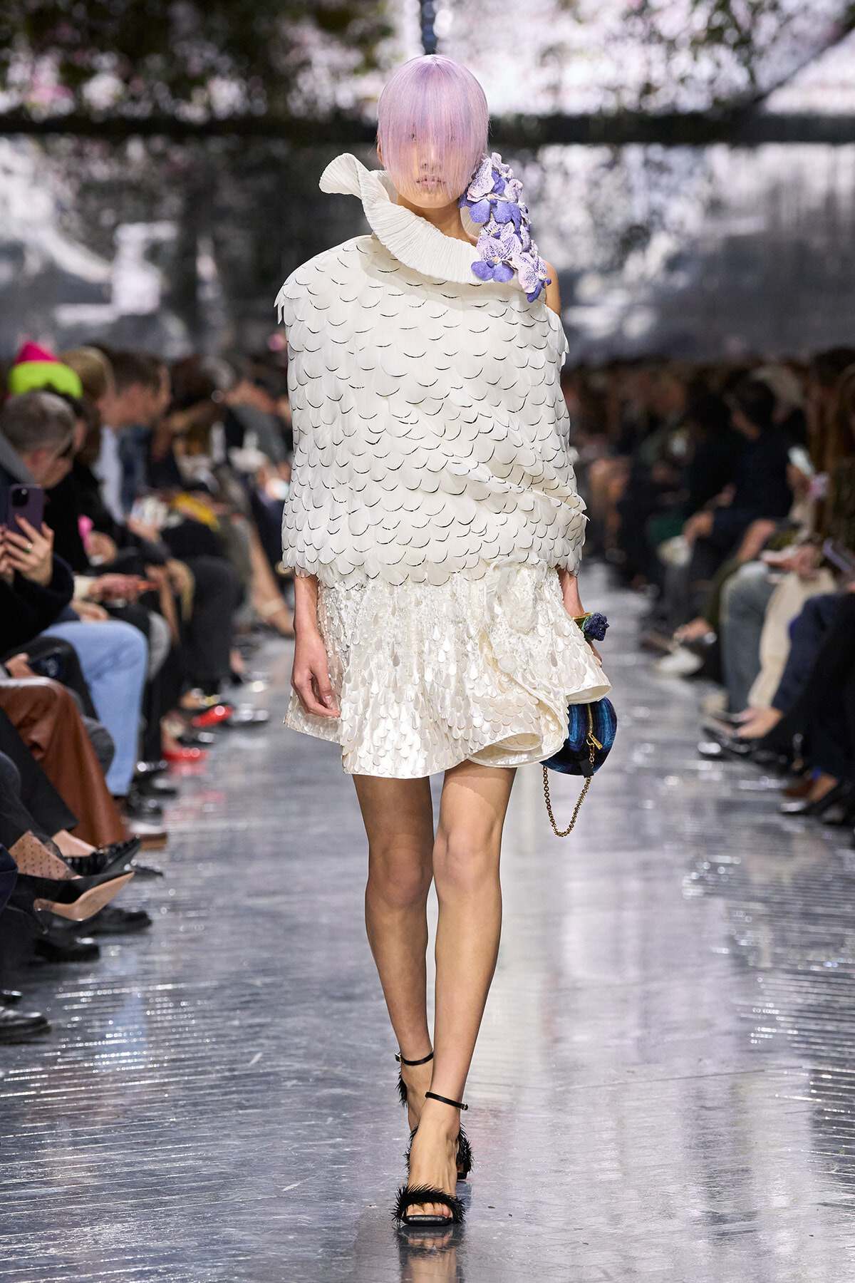 Runway model wearing a white scalloped-feathered cape and matching textured skirt, lavender hair, a purple floral shoulder accent, and a small blue bag with a chain.