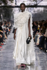 Model on a fashion runway wearing an ivory embroidered gown with a feathered cape, carrying a black floral clutch.