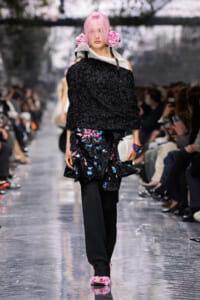 Model with pink hair walking a fashion show in a black textured off-shoulder top and a colorful floral-printed skirt, holding a white clutch.