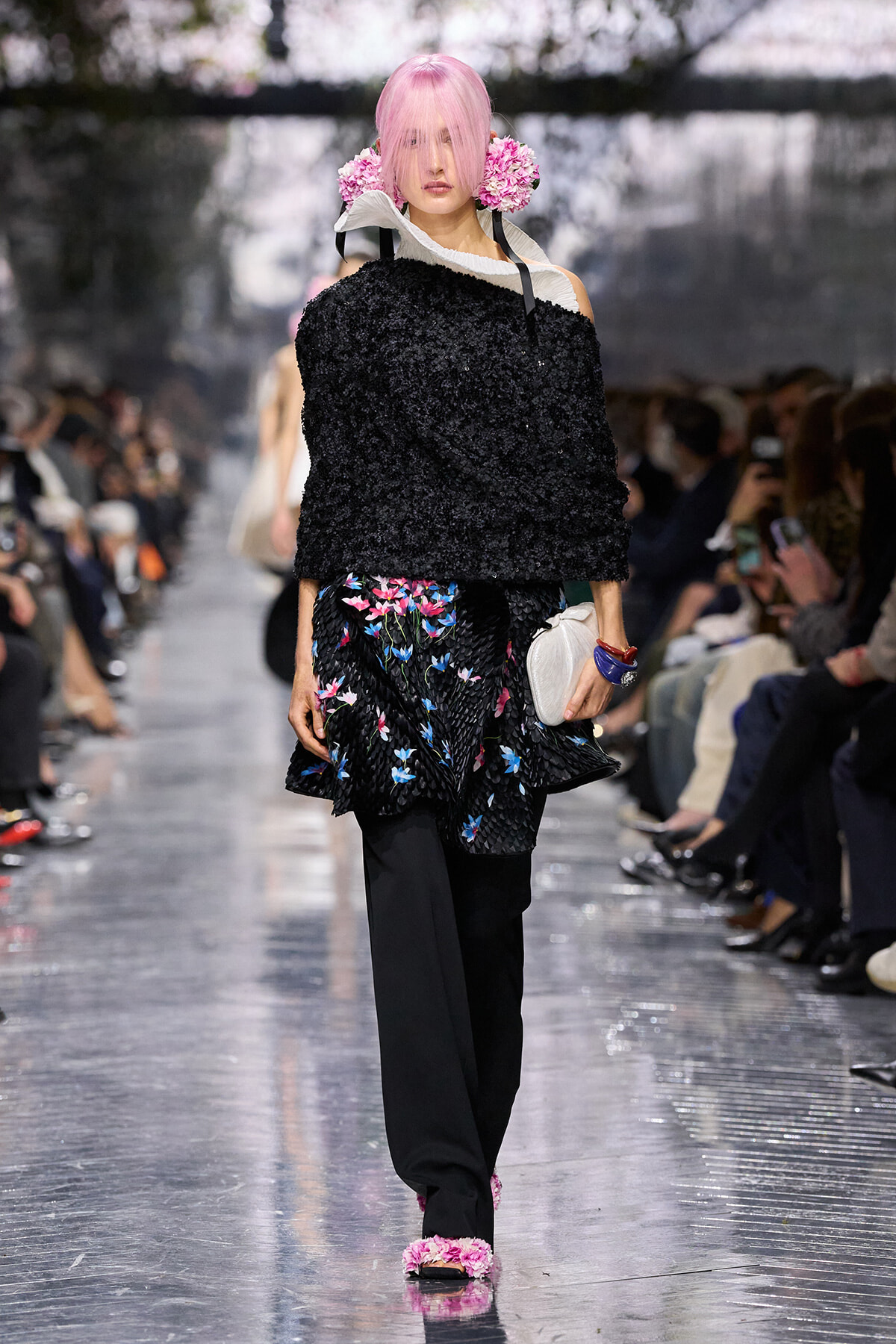 Model with pink hair walking a fashion show in a black textured off-shoulder top and a colorful floral-printed skirt, holding a white clutch.