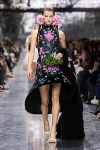 Model walking a fashion-show runway in a black floral-embroidered dress with pink flowers, pink flower hairpieces, and white feathered sandals, holding a green leaf bouquet.
