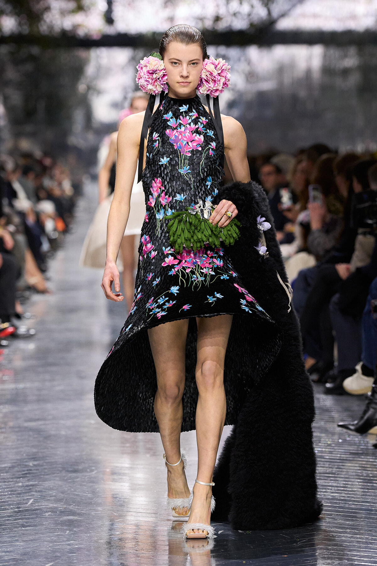 Model walking a fashion-show runway in a black floral-embroidered dress with pink flowers, pink flower hairpieces, and white feathered sandals, holding a green leaf bouquet.