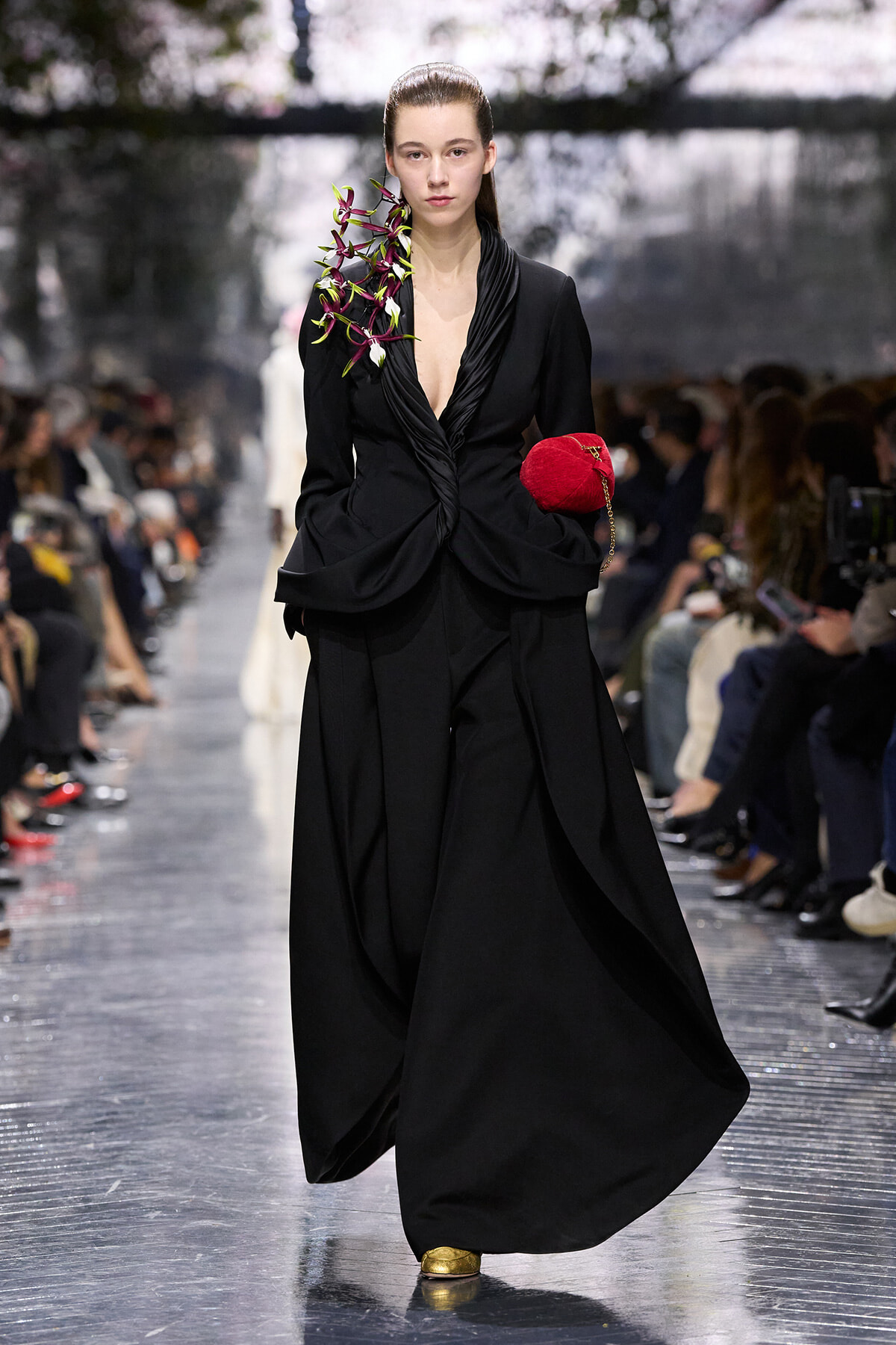 Runway model in a black sculptural gown with an orchid bouquet on her shoulder, carrying a red heart-shaped clutch.