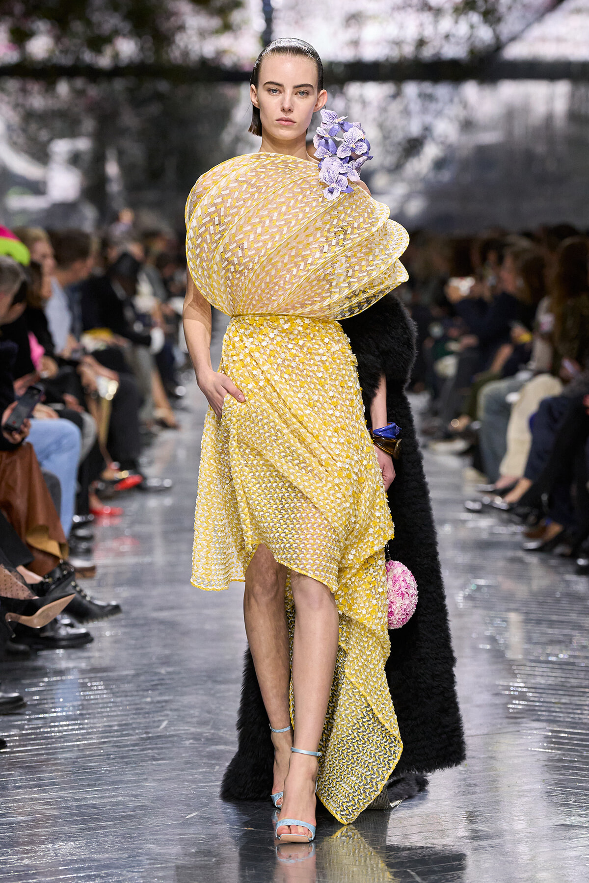 Model walks a fashion runway in a bright yellow, textured dress with a large, sculptural wrap over one shoulder and a cluster of purple flowers at the neckline; blue strappy heels complete the look as spectators watch.