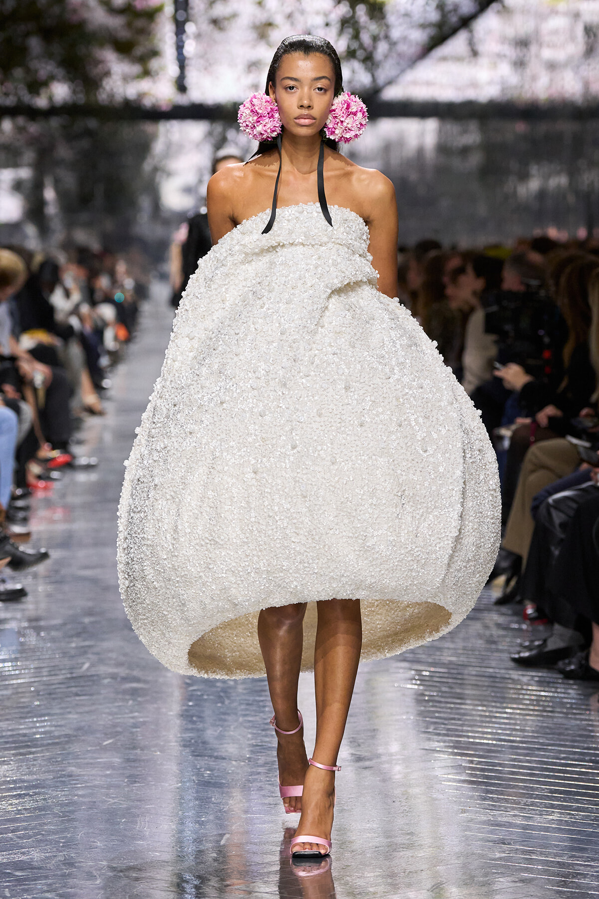 Female model walks a fashion runway wearing a voluminous white textured strapless gown, pink floral earrings, and pink ankle-strap sandals, with an audience in the background.