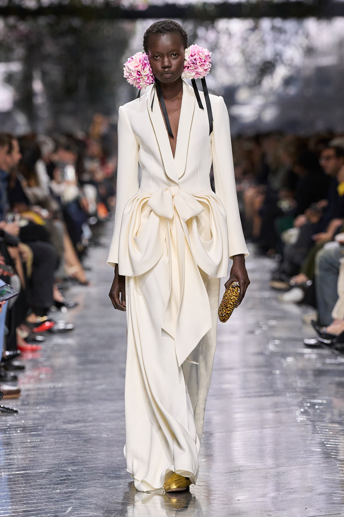 Model on a runway wearing a cream-colored, tailored gown with an oversized bow at the waist, pink flower headpieces, and gold shoes, clutch in hand