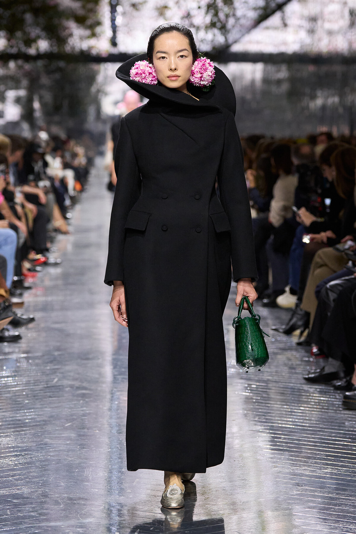 Model walks a runway in a long black double-breasted coat with a sculpted collar, pink flower accessories, and a green crocodile-texture handbag.