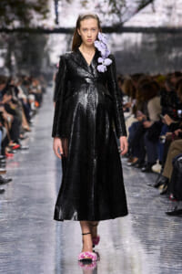 Model on a fashion runway wearing a long black snakeskin coat with a purple flower accessory on the right shoulder and pink floral sandals.