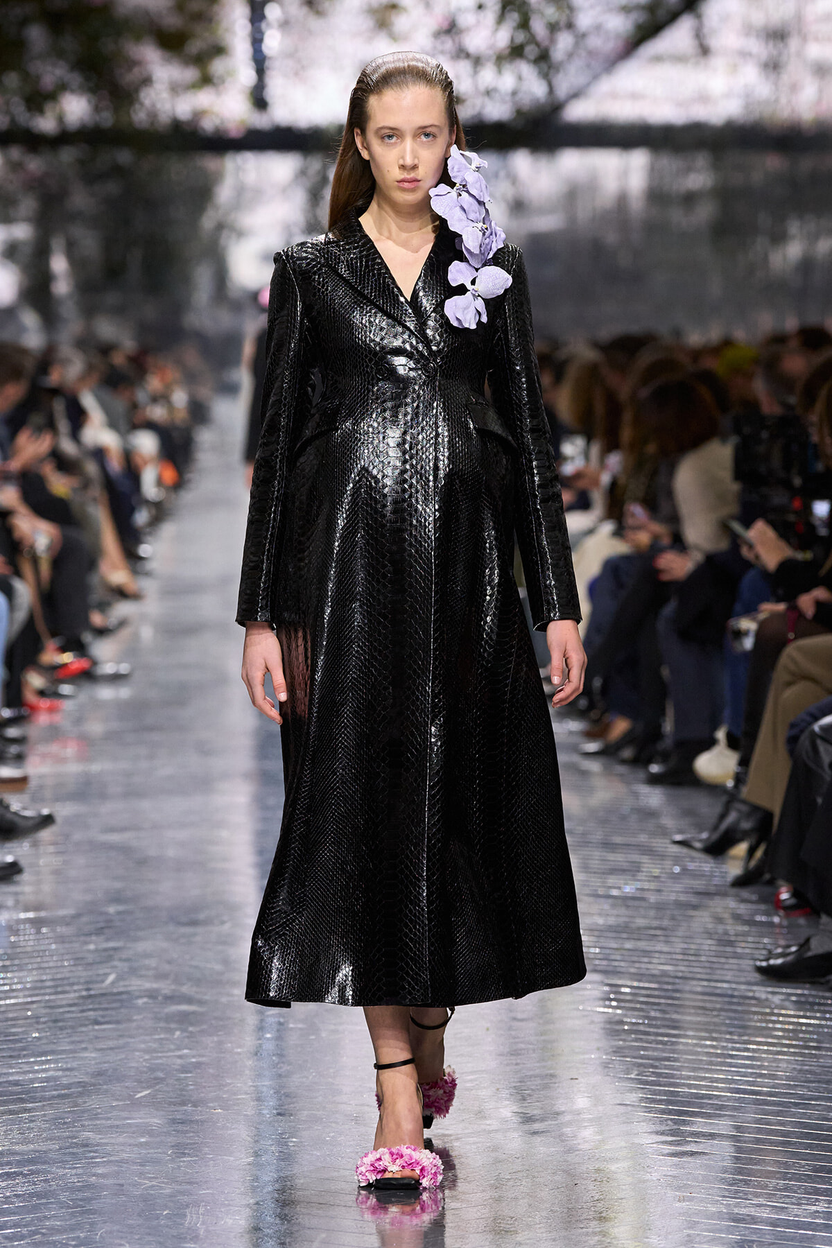 Model on a fashion runway wearing a long black snakeskin coat with a purple flower accessory on the right shoulder and pink floral sandals.