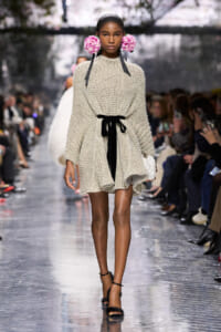 Model walks the runway in a chunky beige knit sweater dress with a black ribbon belt, pink flower ear accessories, and strappy heels on a reflective catwalk.