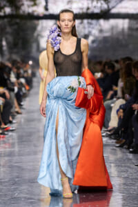 Model on the runway wearing a sheer black top, a light blue embroidered skirt with a slit, and an orange coat draped over her arm, with purple flowers on her shoulder.