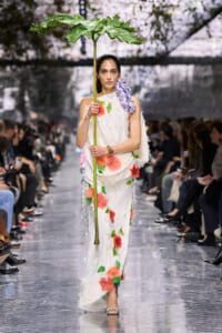 Fashion model on a runway wearing a white floral gown with red flowers and green leaves, holding a tall stem with a large green leaf like a parasol accessory.