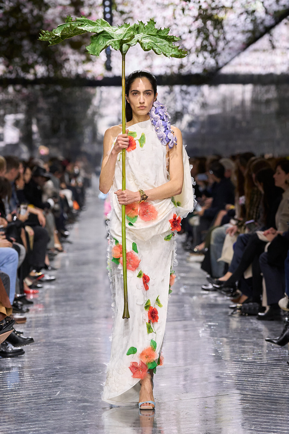 Fashion model on a runway wearing a white floral gown with red flowers and green leaves, holding a tall stem with a large green leaf like a parasol accessory.