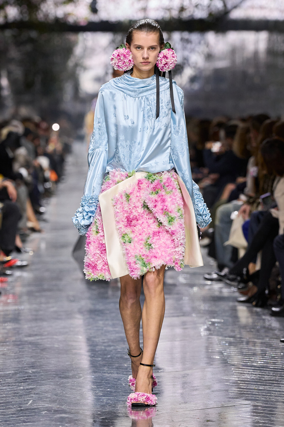 Model walking a fashion runway in a pale blue satin blouse with floral embroidery and a pink floral-textured skirt, wearing pink flower adornments by her ears.