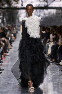Model walks the runway in a white feathered bodice and black layered tulle skirt; audience blurred in the background.