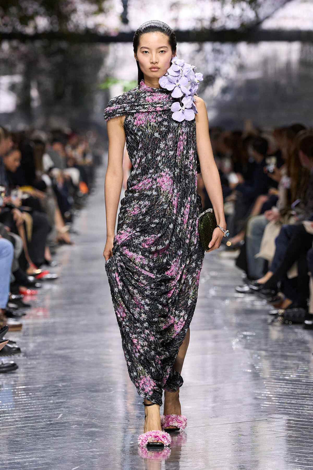 Model walking a dark floral gown with pink accents, oversized purple flowers at the right shoulder, on a glossy runway.