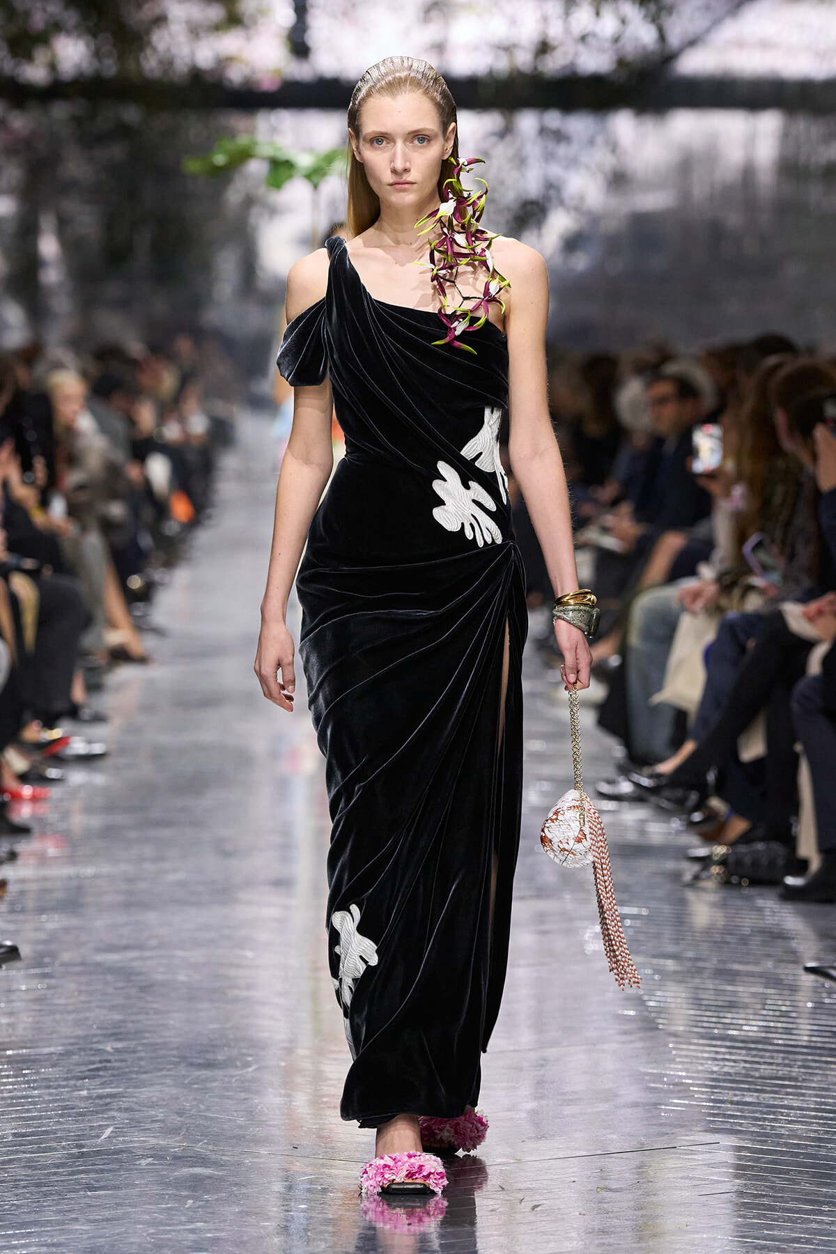 Model on a fashion runway wearing a black velvet one-shoulder gown with white floral appliqués, pink sandals, and a beaded pink handbag.