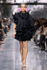 Model walking a runway in a voluminous black ruffled dress with pink flower ear accents.