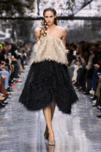 Model walking a runway in a strapless beige feathered top and layered black feather skirt with a purple orchid on her shoulder, audience in background.