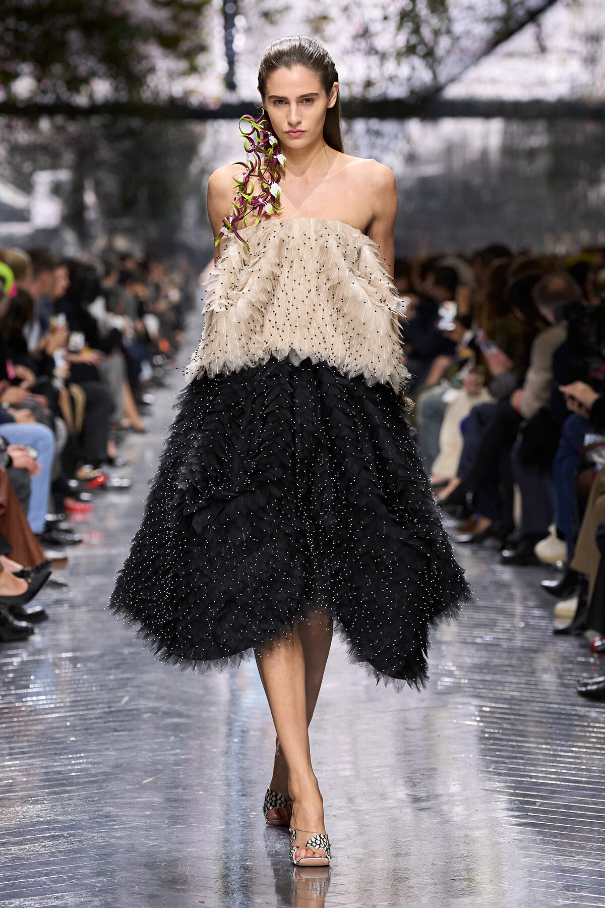 Model walking a runway in a strapless beige feathered top and layered black feather skirt with a purple orchid on her shoulder, audience in background.