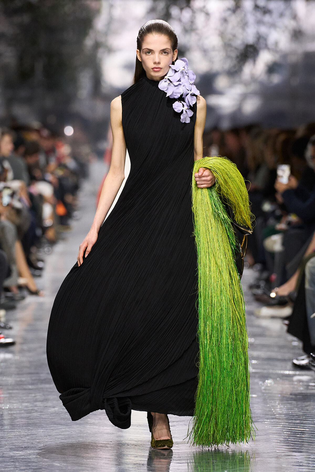 Model on a runway wearing a black pleated gown with lavender floral shoulder detail, holding bright green fringe accessory.