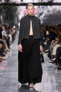 Model on a fashion show runway wearing a textured black cape with an open front, wide-leg black pants, pink flowers tucked behind her ears, and carrying a small black handbag.