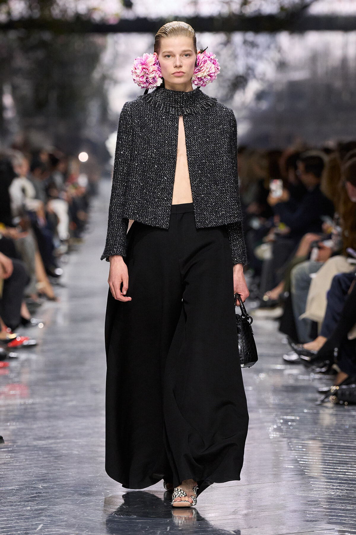 Model on a fashion show runway wearing a textured black cape with an open front, wide-leg black pants, pink flowers tucked behind her ears, and carrying a small black handbag.