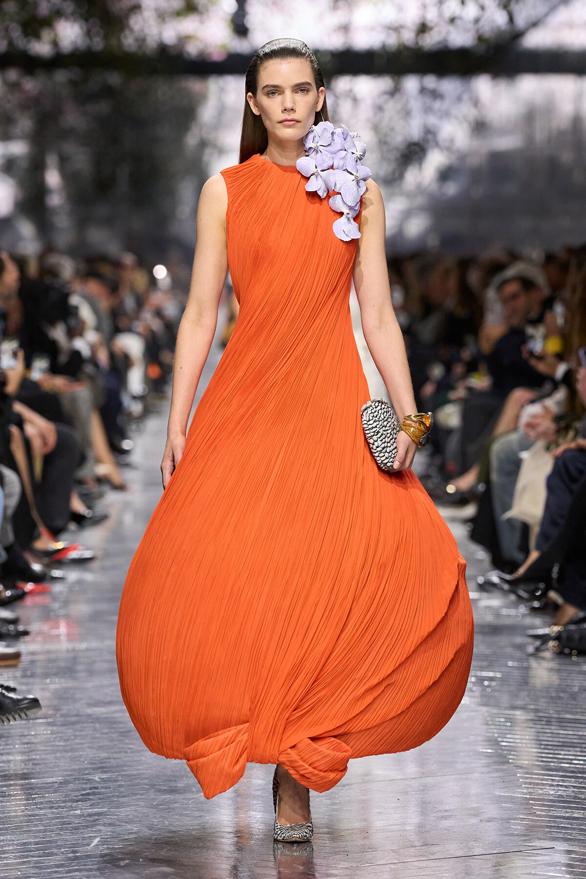 Model walks a runway in a bright orange pleated gown with lilac floral accents at the shoulder, holding a small patterned clutch.