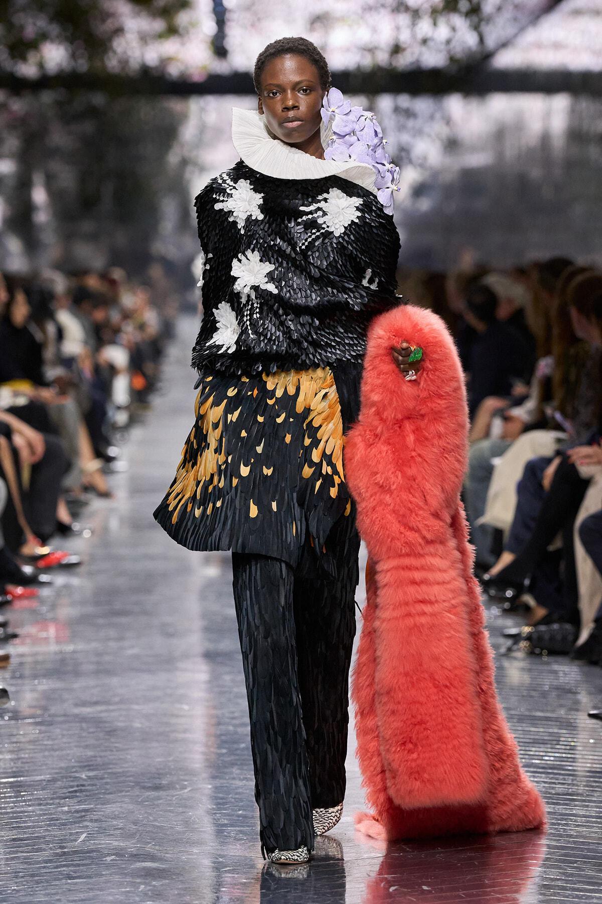 Model walks a fashion runway in a black feathered top with white floral appliqués, a layered yellow‑black feather skirt, a large white ruff collar, lavender flower accessory, and a long pink fluffy coat held at her side.