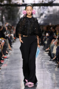Model walks a fashion runway in a black crocheted cape and wide-leg trousers, with pink花 flowers in her hair and a light blue clutch in one hand.