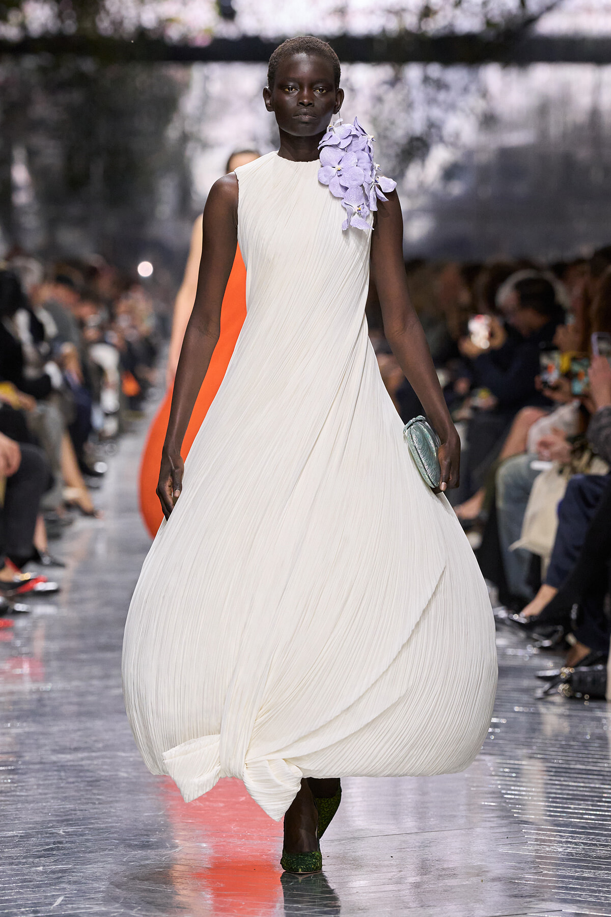 Model on a runway wearing a white pleated gown with a lavender floral shoulder detail, green shoes, and a clutch.