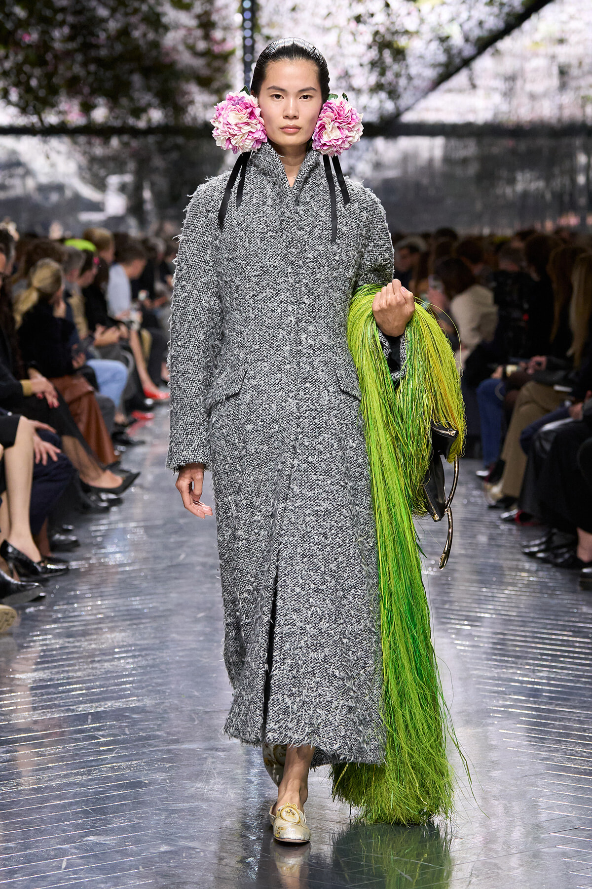 Model on a runway wearing a long gray textured coat with large pink flower ear adornments and a bright green fringe draped from her arm.