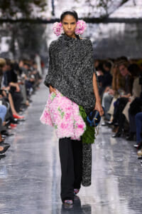 Model walking a fashion runway in a black-and-white textured poncho, pink floral accessories in her hair, a pink floral skirt with green leaves, and a blue clutch appears at her side.