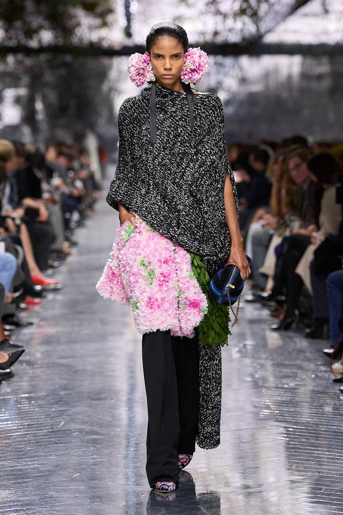 Model walking a fashion runway in a black-and-white textured poncho, pink floral accessories in her hair, a pink floral skirt with green leaves, and a blue clutch appears at her side.
