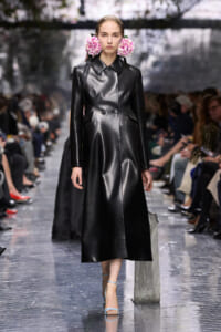 Model wearing a knee-length black leather trench with pink flower accessories, walking the runway.