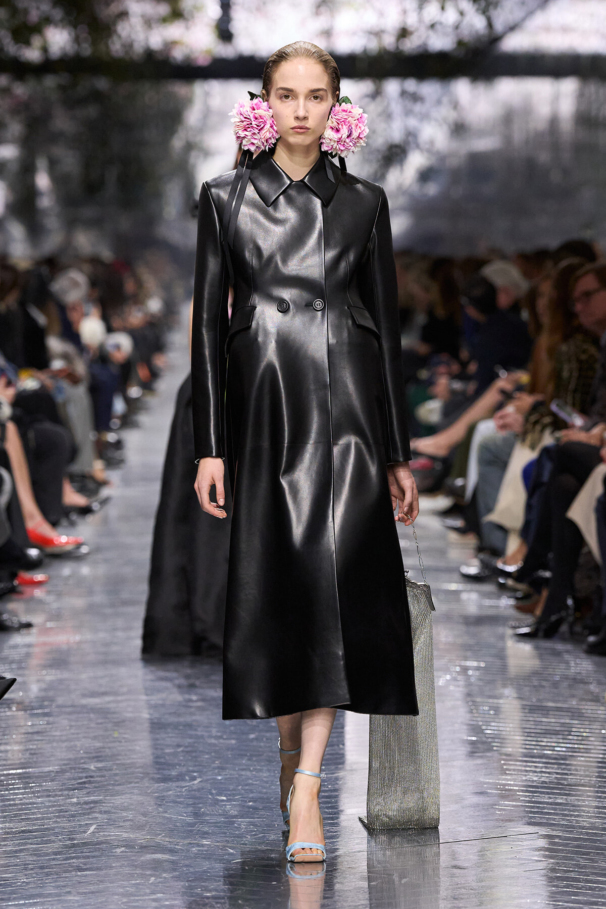 Model wearing a knee-length black leather trench with pink flower accessories, walking the runway.