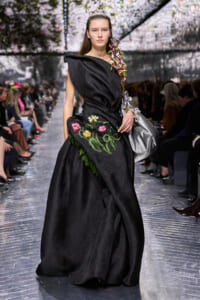 Model walking a runway in a black, sculpted gown with colorful floral embroidery and an orchid on the shoulder.