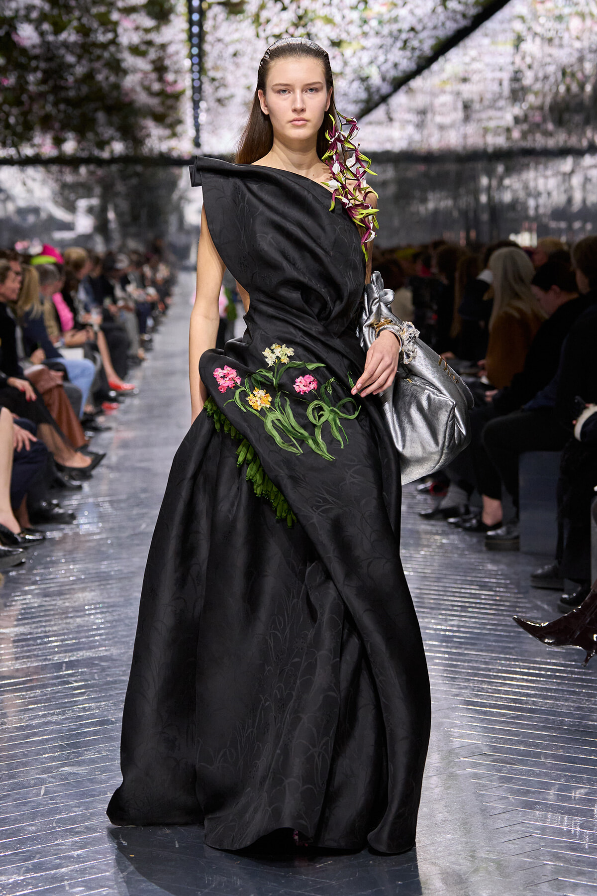 Model walking a runway in a black, sculpted gown with colorful floral embroidery and an orchid on the shoulder.