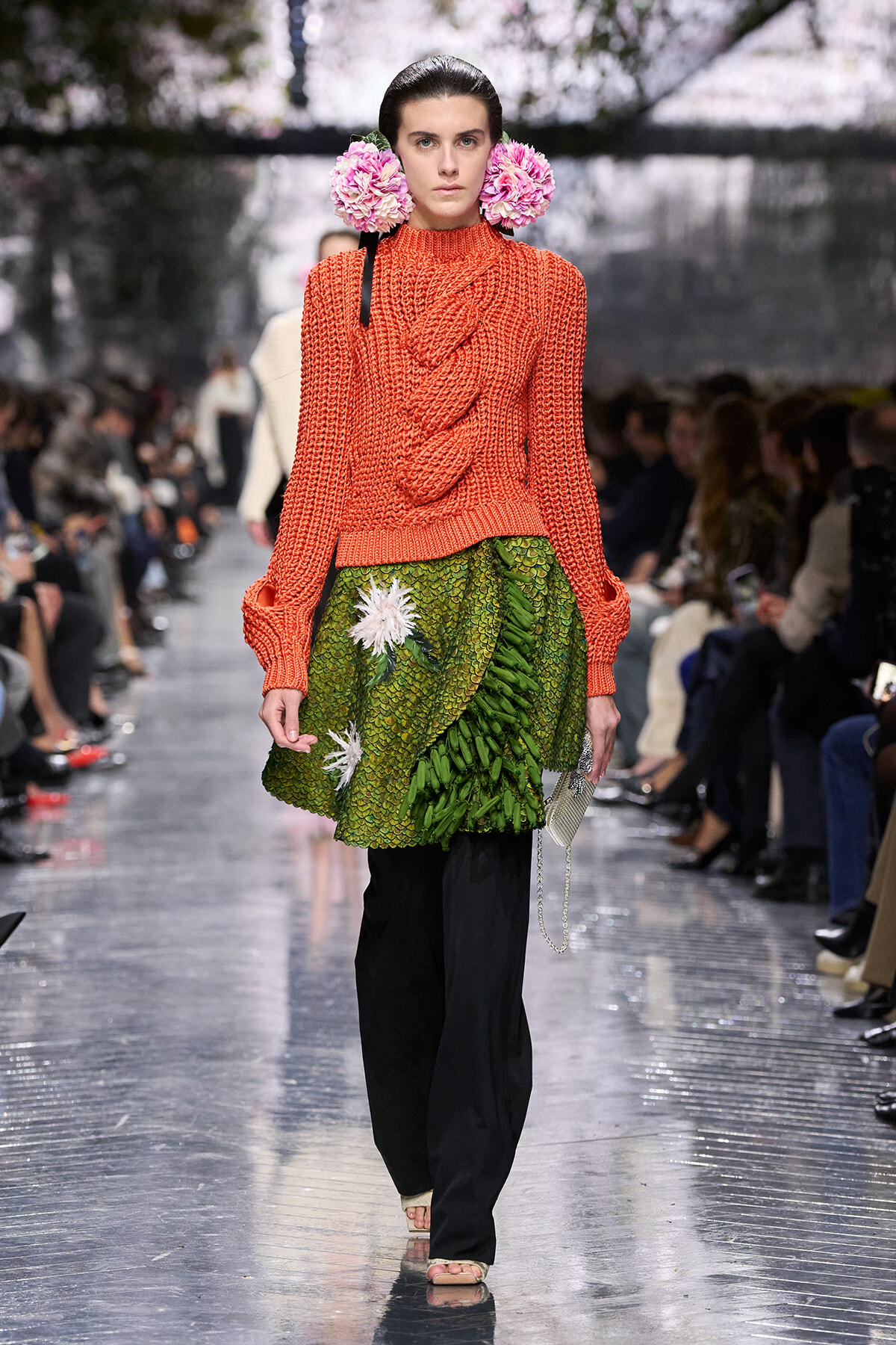 Runway model in a bright orange chunky knit sweater paired with a green leaf-textured skirt and pink floral headpieces at a fashion show.