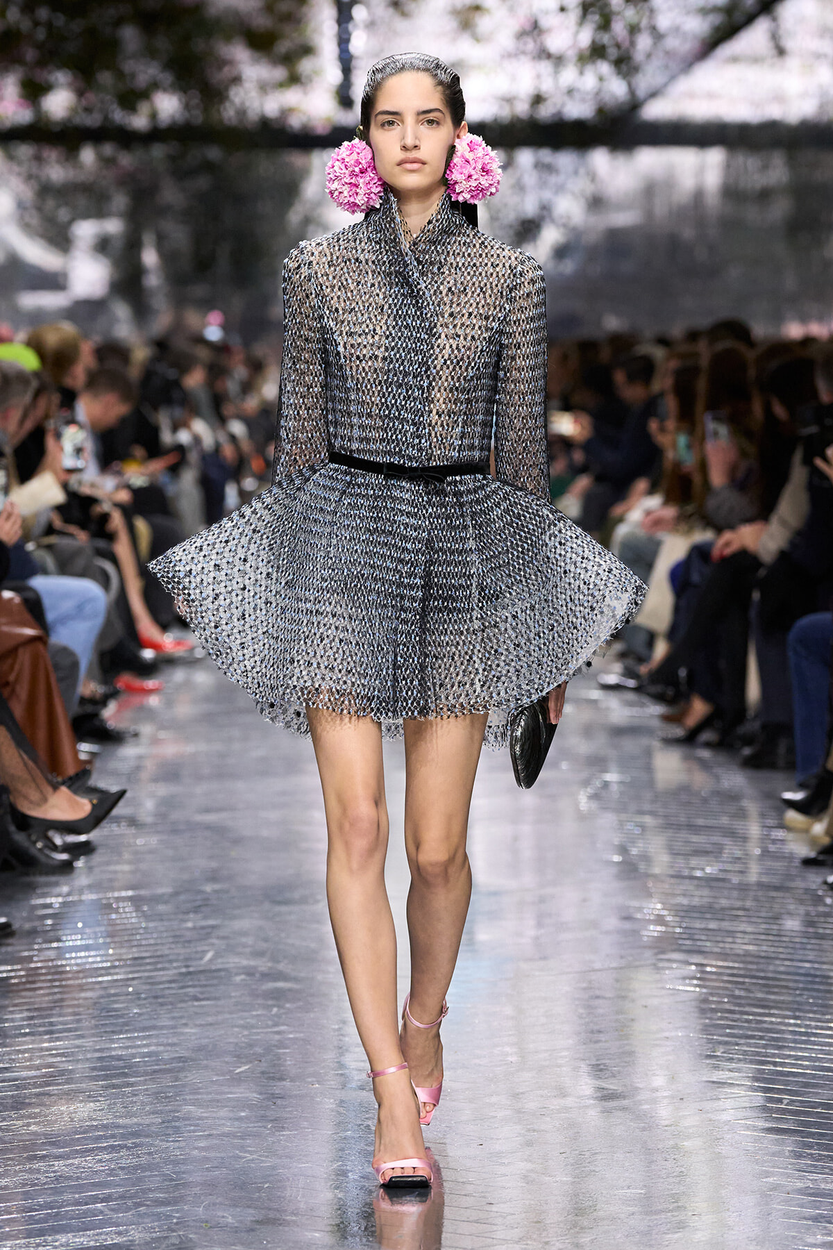 Model walking a runway in a silver textured dress with a structured bodice and a voluminous, flared skirt, wearing pink flower earrings and pink strappy heels.