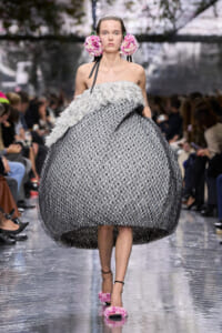 Model walks a couture show wearing an oversized textured ball gown with a white ruffled top and a pink flower headpiece, pink floral sandals.
