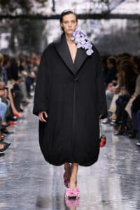 Model walks a runway in an oversized black coat with a lavender orchid bouquet on the left shoulder.