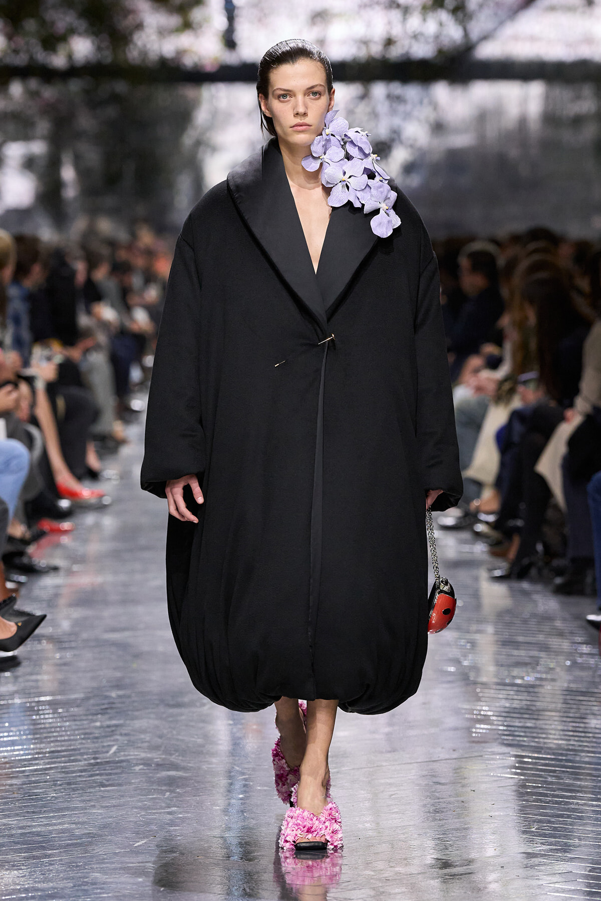 Model walks a runway in an oversized black coat with a lavender orchid bouquet on the left shoulder.