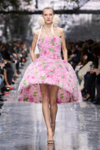 Runway model wearing a pink, flower-covered strapless ball gown with white ribbon straps and big white floral earrings.