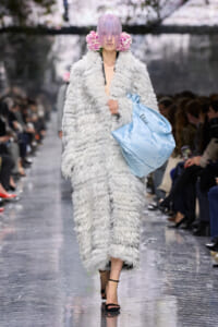 Model struts a voluminous gray faux-fur coat, lavender hair, pink flower accessories, and a large light-blue handbag on a fashion runway.