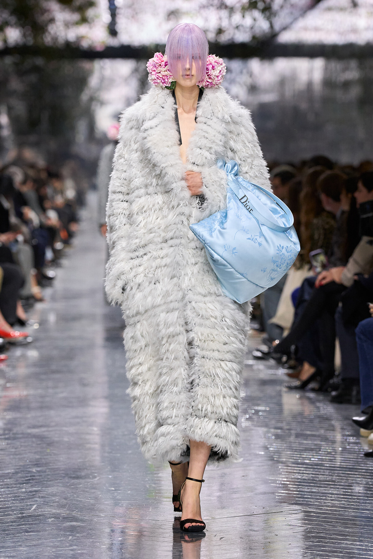 Model struts a voluminous gray faux-fur coat, lavender hair, pink flower accessories, and a large light-blue handbag on a fashion runway.