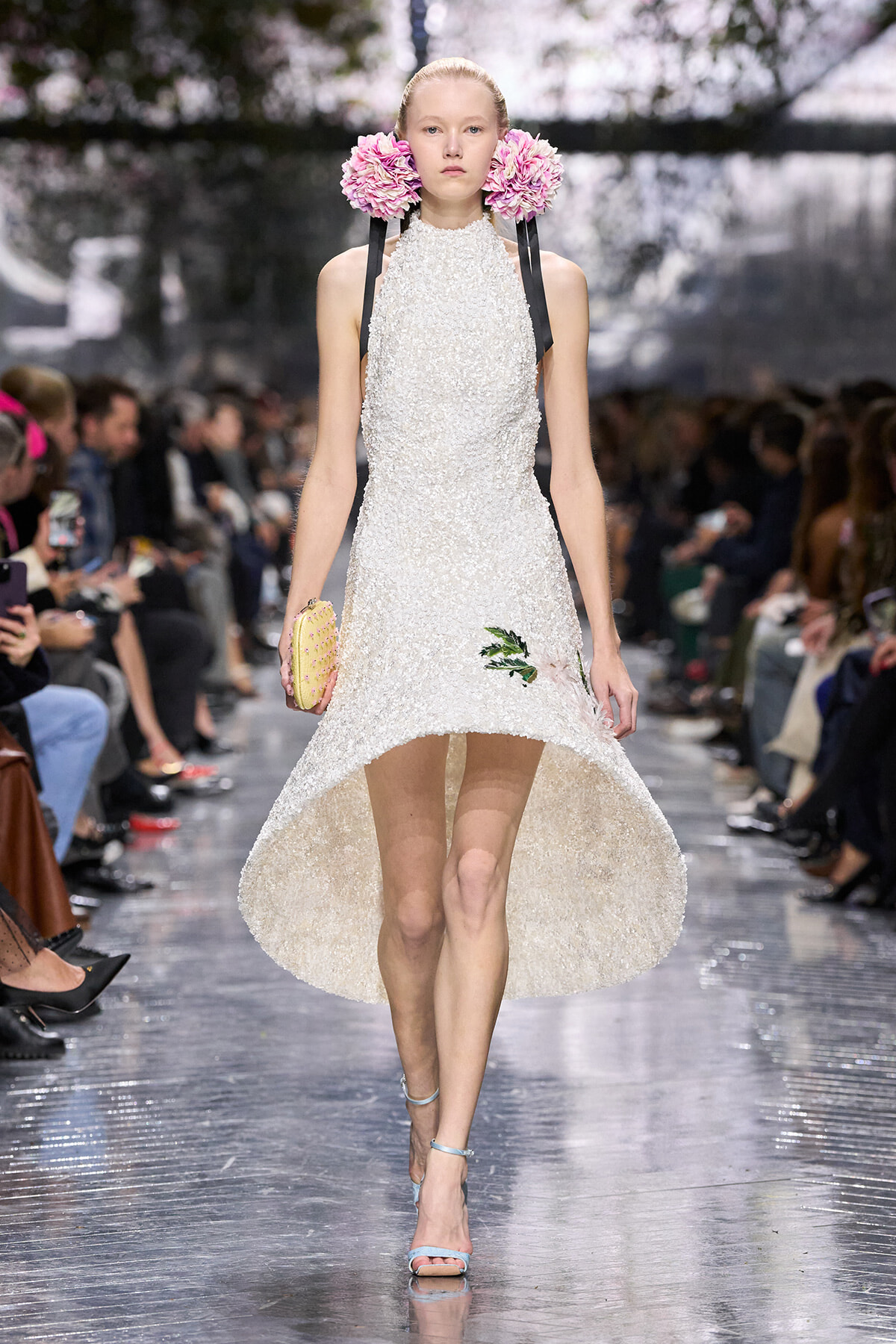 Model walks a runway in a white textured high-low dress with pink flower ear accessories and a pale yellow clutch.