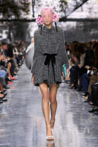 Model walks the runway in a chunky gray knit dress with a black ribbon belt and pink flower headpiece, holding a turquoise clutch.